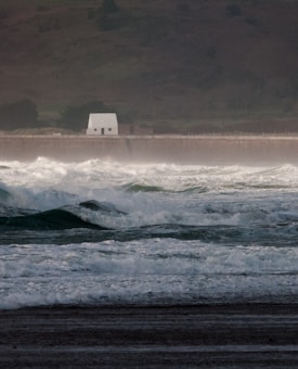 Rough sea waves crash towards a distant shoreline, where a solitary white building stands against a backdrop of dark, rugged hills. The scene appears misty, suggesting turbulent weather conditions.