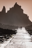 A misty pathway leads through rugged terrain with towering rock formations silhouetted against a dusky sky. Two figures walk side by side along the path, creating a sense of companionship. The surface of the path reflects light, highlighting the texture and wetness of the ground.