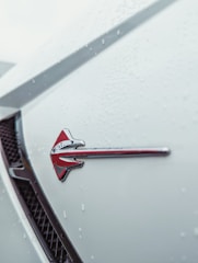 Close-up view of a car's exterior surface covered with raindrops. A red and silver arrow emblem is prominently displayed on the vehicle's white body, with a black grille visible in the background.