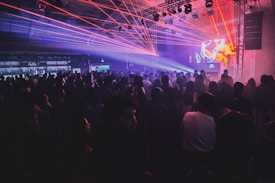 A crowded nightclub or concert venue filled with people standing and moving to music. Red and blue laser lights create a dynamic and energetic atmosphere. A DJ booth is visible on stage with abstract visuals displayed on screens.