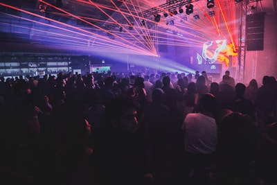 A crowded nightclub or concert venue filled with people standing and moving to music. Red and blue laser lights create a dynamic and energetic atmosphere. A DJ booth is visible on stage with abstract visuals displayed on screens.