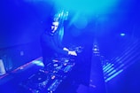A DJ is performing in a dimly lit space filled with blue lighting. The setting appears to be a nightclub or music venue. The DJ is surrounded by DJ equipment, including mixers and turntables. The lighting creates a vibrant, electric atmosphere.