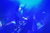 A DJ is performing in a dimly lit space filled with blue lighting. The setting appears to be a nightclub or music venue. The DJ is surrounded by DJ equipment, including mixers and turntables. The lighting creates a vibrant, electric atmosphere.