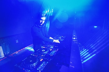 A DJ is performing in a dimly lit space filled with blue lighting. The setting appears to be a nightclub or music venue. The DJ is surrounded by DJ equipment, including mixers and turntables. The lighting creates a vibrant, electric atmosphere.