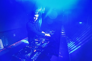 A DJ is performing in a dimly lit space filled with blue lighting. The setting appears to be a nightclub or music venue. The DJ is surrounded by DJ equipment, including mixers and turntables. The lighting creates a vibrant, electric atmosphere.