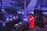 A DJ stands on a stage in a large indoor venue, controlling a set of mixing equipment. He faces a large crowd of people who are illuminated by various colored lights and beams. The atmosphere is lively with bright, dynamic lighting, including blue and purple hues.