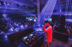 A DJ stands on a stage in a large indoor venue, controlling a set of mixing equipment. He faces a large crowd of people who are illuminated by various colored lights and beams. The atmosphere is lively with bright, dynamic lighting, including blue and purple hues.