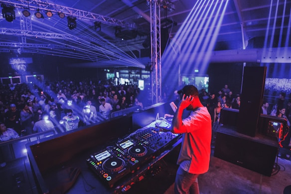 A DJ stands on a stage in a large indoor venue, controlling a set of mixing equipment. He faces a large crowd of people who are illuminated by various colored lights and beams. The atmosphere is lively with bright, dynamic lighting, including blue and purple hues.