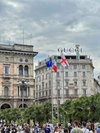 A busy urban scene with a crowd of people gathered in front of historic buildings. The architecture features classical design with ornate facades and arched windows. There are palm trees lining the area, and three flags are raised on poles, including the European Union and Italian flags. The Gucci logo is prominently displayed on top of one of the buildings. The sky is overcast, adding a gray tone to the atmosphere.