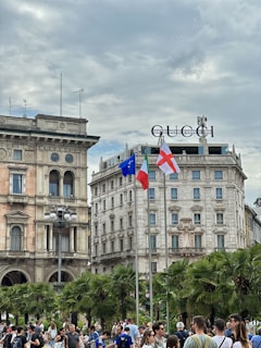 A busy urban scene with a crowd of people gathered in front of historic buildings. The architecture features classical design with ornate facades and arched windows. There are palm trees lining the area, and three flags are raised on poles, including the European Union and Italian flags. The Gucci logo is prominently displayed on top of one of the buildings. The sky is overcast, adding a gray tone to the atmosphere.