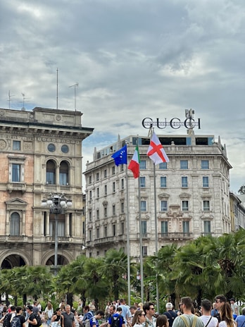 A busy urban scene with a crowd of people gathered in front of historic buildings. The architecture features classical design with ornate facades and arched windows. There are palm trees lining the area, and three flags are raised on poles, including the European Union and Italian flags. The Gucci logo is prominently displayed on top of one of the buildings. The sky is overcast, adding a gray tone to the atmosphere.