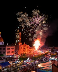 A lively nighttime scene depicts a festive event with a large crowd gathered around a church. Fireworks light up the sky above the church, which is adorned with colorful lights and decorations. Market stalls and bright tents are visible, suggesting a street fair or carnival atmosphere.