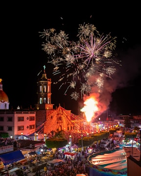 A lively nighttime scene depicts a festive event with a large crowd gathered around a church. Fireworks light up the sky above the church, which is adorned with colorful lights and decorations. Market stalls and bright tents are visible, suggesting a street fair or carnival atmosphere.