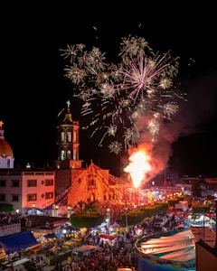 A lively nighttime scene depicts a festive event with a large crowd gathered around a church. Fireworks light up the sky above the church, which is adorned with colorful lights and decorations. Market stalls and bright tents are visible, suggesting a street fair or carnival atmosphere.