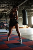 A person is practicing boxing in a gym setting, standing in a fighting stance on red and blue puzzle mats. The gym is dimly lit with a large punching bag hanging in the background. The individual is wearing boxing gloves and athletic wear, and appears focused and ready.