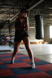 A person is practicing boxing in a gym setting, standing in a fighting stance on red and blue puzzle mats. The gym is dimly lit with a large punching bag hanging in the background. The individual is wearing boxing gloves and athletic wear, and appears focused and ready.