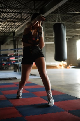 A person is practicing boxing in a gym setting, standing in a fighting stance on red and blue puzzle mats. The gym is dimly lit with a large punching bag hanging in the background. The individual is wearing boxing gloves and athletic wear, and appears focused and ready.