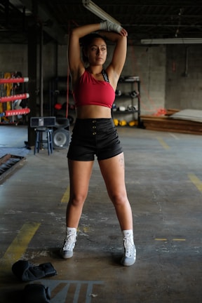A person is standing in a boxing gym, stretching with arms raised above their head. The individual is wearing a red sports top, black shorts, and white athletic shoes. Boxing gloves are on the floor nearby, and gym equipment can be seen in the background.