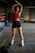 A person is standing in a boxing gym, stretching with arms raised above their head. The individual is wearing a red sports top, black shorts, and white athletic shoes. Boxing gloves are on the floor nearby, and gym equipment can be seen in the background.