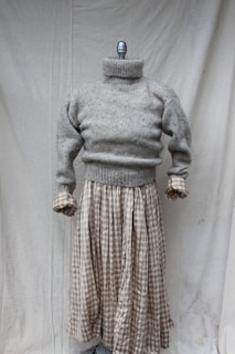 A mannequin displaying a gray knitted turtleneck sweater paired with a long plaid skirt. The setting is minimal with a plain fabric backdrop.