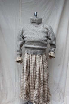 A mannequin displaying a gray knitted turtleneck sweater paired with a long plaid skirt. The setting is minimal with a plain fabric backdrop.