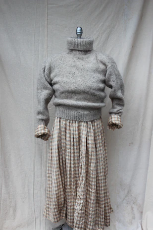 A mannequin displaying a gray knitted turtleneck sweater paired with a long plaid skirt. The setting is minimal with a plain fabric backdrop.