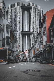 A modern, high-rise building towers over a narrow street lined with older, colorful shophouses. The perspective emphasizes the height and unique architecture of the structure. Cars and people are present on the street, and numerous signs hang from the buildings, adding to the urban atmosphere.