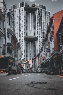A modern, high-rise building towers over a narrow street lined with older, colorful shophouses. The perspective emphasizes the height and unique architecture of the structure. Cars and people are present on the street, and numerous signs hang from the buildings, adding to the urban atmosphere.
