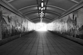 A long, narrow tunnel with an arched ceiling is lined with tiled walls, featuring intricate murals. The tunnel extends towards a bright light at its end, creating a sense of depth and perspective. The flooring consists of large square tiles, and overhead lights run the length of the ceiling, casting a soft glow.