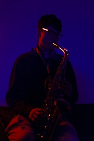 A person is playing a saxophone in a dimly lit, moody environment. The lighting casts a deep blue and red glow, highlighting the contours of the instrument and the silhouette of the musician. The atmosphere feels intimate and introspective.
