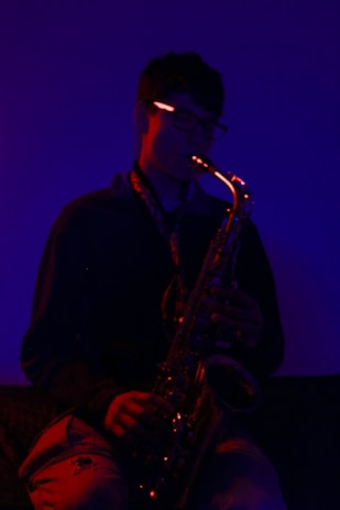 A person is playing a saxophone in a dimly lit, moody environment. The lighting casts a deep blue and red glow, highlighting the contours of the instrument and the silhouette of the musician. The atmosphere feels intimate and introspective.