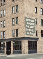 A brick building corner with multiple windows features a vintage sign advertising 'Modern Rooms To Rent by Week or Month.' The structure is on a city street with a concrete sidewalk, a trash can, and a bench nearby.