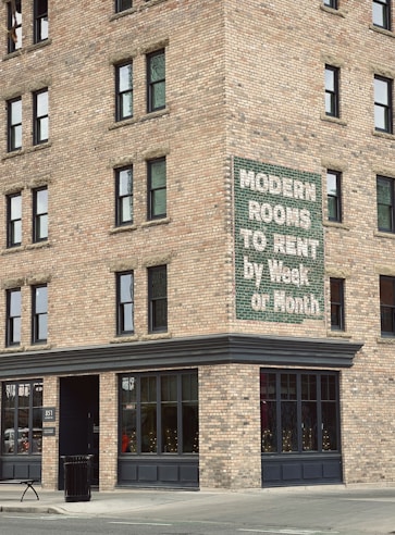 A brick building corner with multiple windows features a vintage sign advertising 'Modern Rooms To Rent by Week or Month.' The structure is on a city street with a concrete sidewalk, a trash can, and a bench nearby.