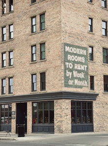 A brick building corner with multiple windows features a vintage sign advertising 'Modern Rooms To Rent by Week or Month.' The structure is on a city street with a concrete sidewalk, a trash can, and a bench nearby.