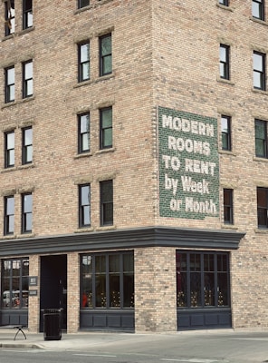 A brick building corner with multiple windows features a vintage sign advertising 'Modern Rooms To Rent by Week or Month.' The structure is on a city street with a concrete sidewalk, a trash can, and a bench nearby.