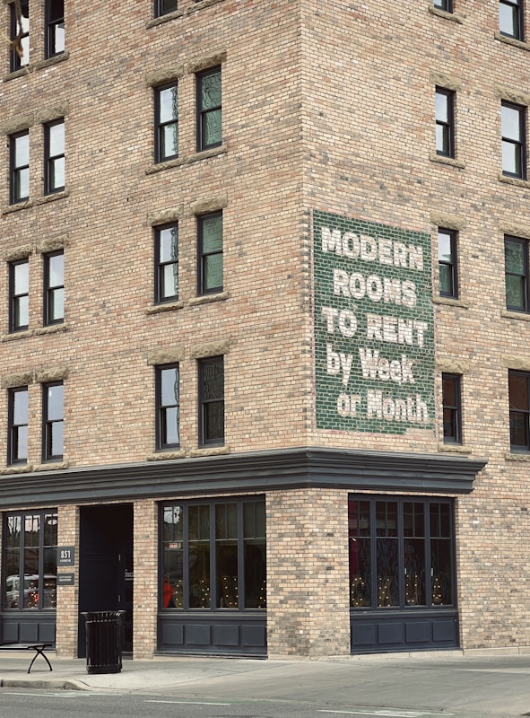 A brick building corner with multiple windows features a vintage sign advertising 'Modern Rooms To Rent by Week or Month.' The structure is on a city street with a concrete sidewalk, a trash can, and a bench nearby.