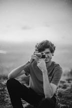 A person is sitting outdoors while holding a vintage camera up to their face, appearing to take a photo. The scene is in black and white, adding a nostalgic feel. Their expression is focused, and the background appears to be blurred natural scenery.