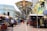 A bustling outdoor market scene with numerous stalls under large decorative metalwork. People walk along a tiled walkway flanked by colorful buildings and shops, some of which display bags and other goods. Street lamps and signage can be seen along the path.