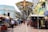 A bustling outdoor market scene with numerous stalls under large decorative metalwork. People walk along a tiled walkway flanked by colorful buildings and shops, some of which display bags and other goods. Street lamps and signage can be seen along the path.