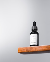 A black dropper bottle labeled 'Minimalist Retinol 0.5%' sits on a wooden shelf against a light gray background. The design is simple and modern, emphasizing the minimalist branding.