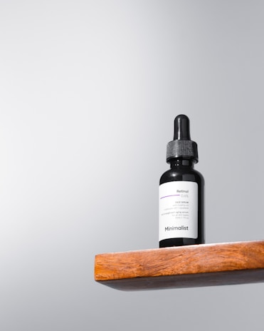 A black dropper bottle labeled 'Minimalist Retinol 0.5%' sits on a wooden shelf against a light gray background. The design is simple and modern, emphasizing the minimalist branding.