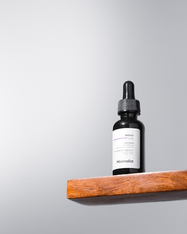 A black dropper bottle labeled 'Minimalist Retinol 0.5%' sits on a wooden shelf against a light gray background. The design is simple and modern, emphasizing the minimalist branding.