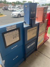 The image shows a row of newspaper vending machines placed on a sidewalk. There are four machines, each differing in color, with visible text and images on their front panels. Some of the text is in Korean. Behind the machines, there is a parking lot with several cars parked and a few people walking.