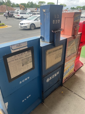 The image shows a row of newspaper vending machines placed on a sidewalk. There are four machines, each differing in color, with visible text and images on their front panels. Some of the text is in Korean. Behind the machines, there is a parking lot with several cars parked and a few people walking.