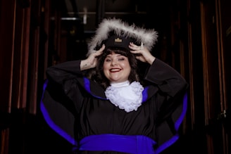 A person is wearing a graduation cap with white feathers and a traditional graduation gown with blue accents and a white ruffled collar, smiling broadly with hands adjusting the cap.