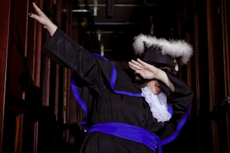 A person dressed in graduation attire performs a dab pose in a dimly lit hallway. The outfit features a black gown with blue accents, white lace, and a cap adorned with white feathers.