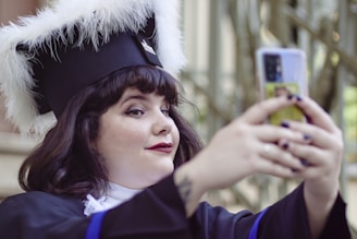 A person wearing a graduation cap with white fur trim is taking a selfie with a smartphone. They have dark hair and are dressed in a graduation gown. The background is blurred, giving prominence to the person in focus.