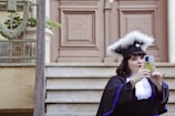 A person wearing a graduation cap with white feathers takes a selfie on a smartphone while sitting on steps in front of an ornate wooden door.