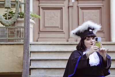 A person wearing a graduation cap with white feathers takes a selfie on a smartphone while sitting on steps in front of an ornate wooden door.