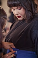 A person in a black outfit is having their blue garment adjusted by another individual. The focus is on the process of fastening or zipping up the clothing, with attention to detail in the clothing and the hands assisting.